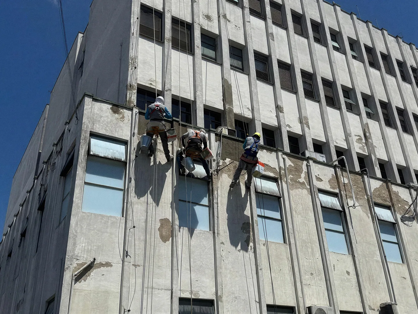 Três pintores trabalhando em altura na fachada desgastada de um prédio comercial